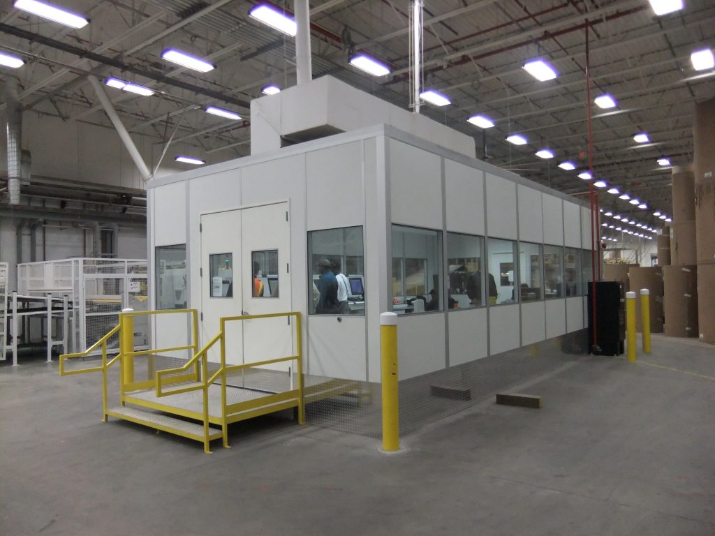 Custom equipment enclosure built by MachineEnclosure.com inside a manufacturing facility, showing modular panels, windows, and double-door access.